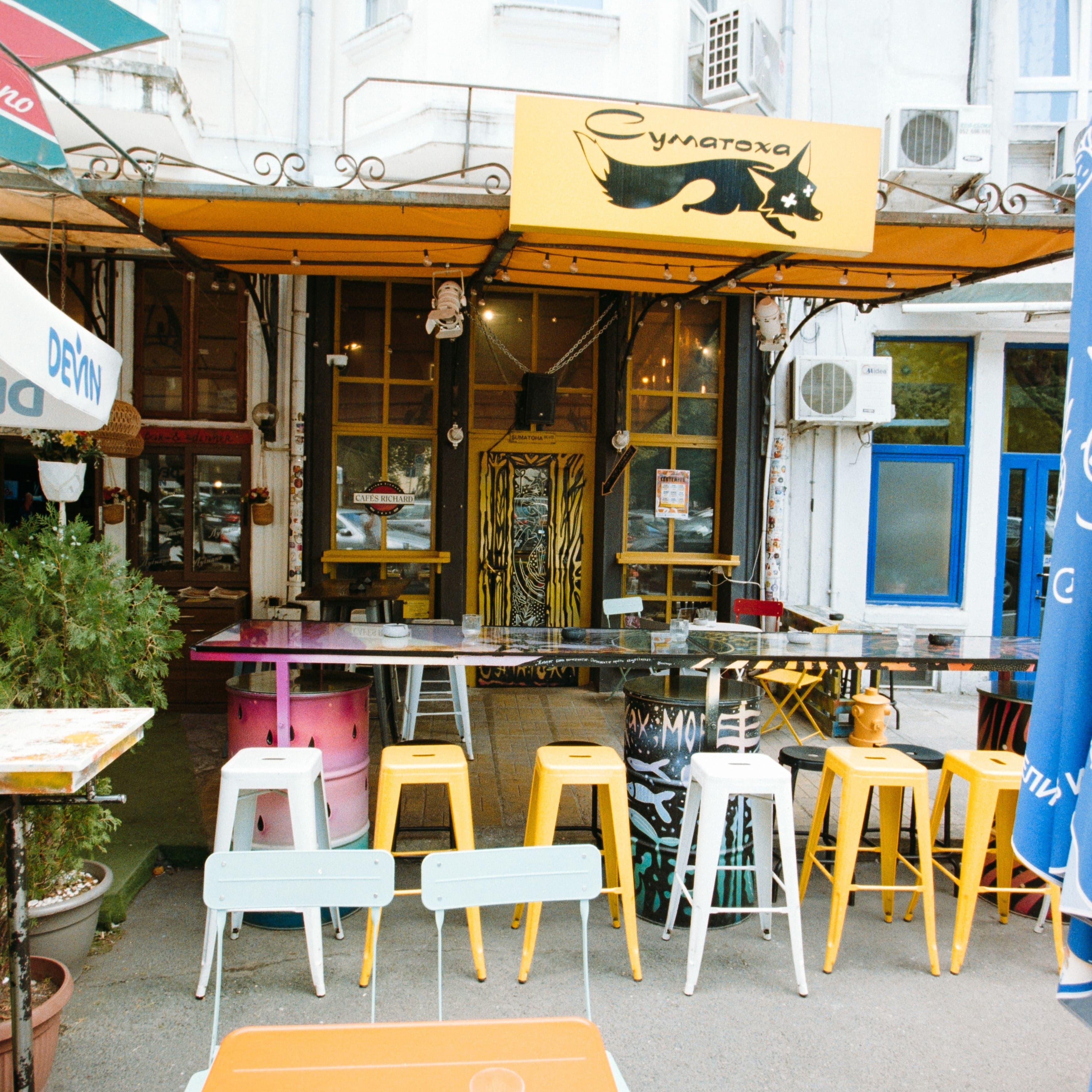 Outdoor seating area with colorful tables and chairs in front of a building with a sign.